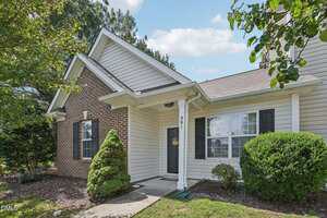 Single-story house with a mix of brick and beige siding exterior, black shutters, a covered entryway with a white column, and trimmed shrubs and plants in the front yard.