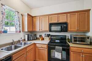 A kitchen with wooden cabinets, a double sink, a black stove with oven, a black microwave, toaster oven, knife block, and various appliances on beige countertops. A window with patterned curtains is above the sink.