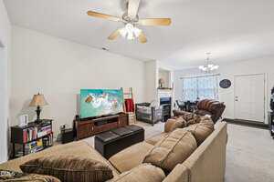 A living room with a beige sectional sofa, a TV displaying an aerial view of a beach, a ceiling fan with lights, a bookcase, a small fireplace, a baby crib, and a dining area with a table and chairs in the background.