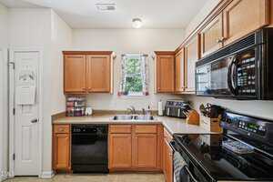 Kitchen with wooden cabinets, black appliances including a stove, microwave, and dishwasher, a double sink under a window with floral curtains, and various kitchen items on the countertops.