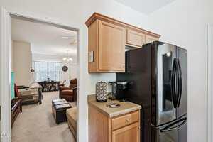A small kitchen area with wooden cabinets, a black refrigerator, a coffee maker, and a pod holder. The view extends into a living room with beige carpet, brown furniture, and a dining area with a chandelier.