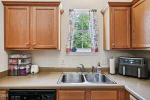 A kitchen countertop with a double sink under a window, surrounded by wooden cabinets. On the left are organized containers and appliances; on the right is a coffee maker. A curtain covers part of the window.