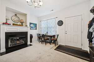A dining area with a rectangular table and six chairs near a window with patterned curtains, a fireplace with a clock and decor on the mantel, and a closed white door with coats hanging nearby.