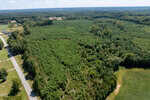 Aerial view of a large, densely wooded area with evergreen trees, bordered by a paved road on the left and open fields. Scattered houses and more forest are visible in the distance under a clear sky.