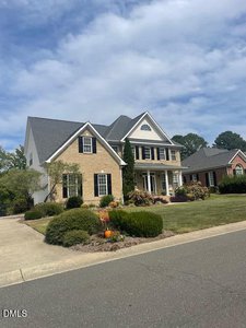 Two-story suburban house with light brick and white siding, a gabled roof, manicured shrubs, and a paved driveway. The lawn is neatly trimmed and the sky is partly cloudy.