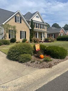 A two-story brick house on 2639 Bowden Drive with black shutters and white trim, surrounded by neatly trimmed bushes and a landscaped front yard in Creedmoor. A small garden bed near the sidewalk features a pumpkin and a colorful flag.