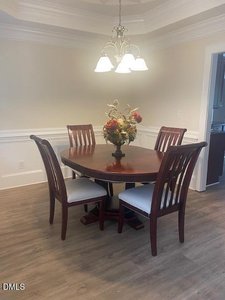 A round wooden dining table with four matching chairs and a floral centerpiece sits under a five-light chandelier in a room with light-colored walls, white trim, and wood-style flooring.