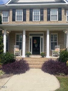 A two-story brick house with black shutters, white columns, and a covered porch. Two white rocking chairs with cushions sit by the front door, which is decorated with a floral wreath. Bushes and plants line the walkway.