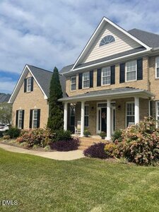 Two-story brick house with black shutters and white trim, featuring a covered front porch with two rocking chairs, surrounded by neatly trimmed bushes and a well-maintained lawn under a partly cloudy sky.