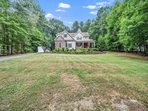 A two-story house with beige siding and brick accents sits at the end of a long driveway, surrounded by tall trees and a large, neatly maintained front lawn under a blue sky with scattered clouds.