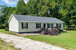 A single-story beige house with black shutters and a dark gray roof sits on a grassy lot. Wooden steps lead to a small porch, and a gravel driveway runs alongside the house. Trees are in the background.