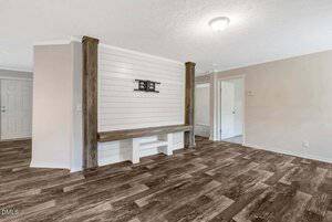 A room with wood-look flooring, light beige walls, a white shiplap accent wall with mounted TV bracket, and built-in wooden bench, adjacent to a hallway and open entryway.