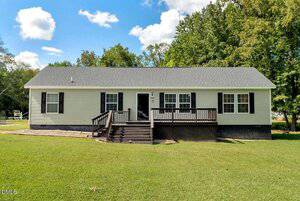 A single-story house at 5766 Raleigh Road, Kittrell, features beige siding, black shutters, and a dark roof. It has a front porch with steps to the lawn, framed by trees and a blue sky with scattered clouds in the background.