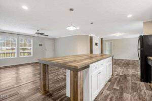 A spacious kitchen and living area with wood-look tile flooring, a large island with a wooden countertop and white cabinets, ceiling lights, a black refrigerator, and windows letting in natural light.
