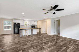Open-plan kitchen and living area with light-colored walls, dark wood-style flooring, a black ceiling fan, a kitchen island with two stools, white cabinets, and a black refrigerator and stove.