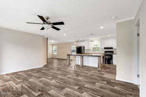 Open-concept living area with wood-look flooring, a ceiling fan, and neutral walls. The kitchen features white cabinets, a large island with seating, and stainless steel appliances. There are large windows letting in natural light.