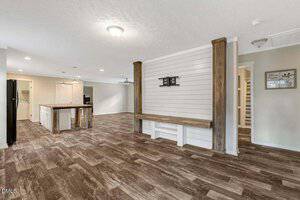 Open-concept living space with wood-look tile flooring, a central kitchen with an island, white cabinetry, and a black refrigerator. The area features white walls, shiplap accents, and exposed wooden beams.