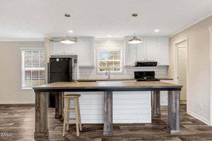 A modern kitchen features a large island with a wooden countertop and one stool, white cabinets, a black refrigerator, a black stove, two pendant lights, and a window above the sink.