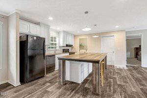 A modern kitchen with wood-look flooring, a black refrigerator, black dishwasher, white cabinets, a center island with a wooden countertop and two stools, and neutral-colored walls. A bedroom is visible through a doorway.