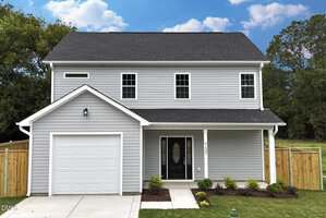 A two-story gray house with white trim at 735 Baker Street, Oxford, featuring a black front door, single-car garage, concrete driveway, small shrubs, and a fenced backyard set against trees and a blue sky with clouds.