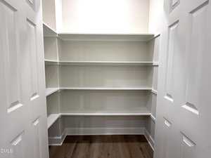 A walk-in pantry with white built-in shelves on three walls, double white doors, and a wood floor. The shelves are empty and the space is well-lit.