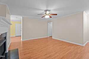 A spacious living room with light beige walls, white trim, a ceiling fan with lights, wood flooring, a white mantel fireplace on the left, and multiple doorways leading to other rooms.