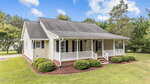 Charming single-story beige house with white trim at 6581 Hwy 158 Bus, Henderson. Features a black shingle roof, covered front porch with white railings, manicured shrubs, lush green grass, and a tree-lined yard.