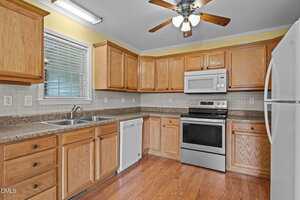 A kitchen with wood cabinets, granite countertops, stainless steel oven and microwave, white refrigerator and dishwasher, double sink under a window, tile backsplash, wood flooring, and a ceiling fan with lights.