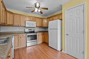 Kitchen with wooden cabinets, white refrigerator, microwave, electric stove, stainless steel sink, brown countertops, and wood-look flooring. Ceiling fan and light fixture overhead, walls painted yellow, door to the right.