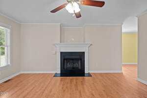 A living room with light beige walls, hardwood floors, a black fireplace with a white mantel, ceiling fan with lights, and a large window with white blinds letting in natural light.