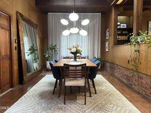 A dining room with a wooden table, six chairs, a modern chandelier, a floral centerpiece, plants, a large mirror on the left wall, sheer curtains, a textured rug, and wood and brick walls.