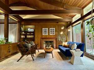 A mid-century modern living room with wood-paneled walls, exposed beams, large windows, a blue sofa, two brown armchairs, a wooden coffee table, a fireplace, bookshelves, and various plants.