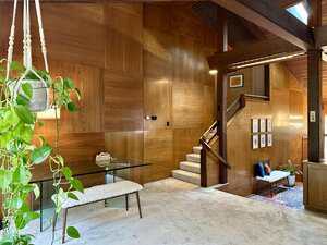 A room with wood-paneled walls, carpeted floors, a glass table with a decorative bowl, a white bench, a hanging plant, framed artwork, and a staircase leading to an upper level. Warm natural light fills the space.