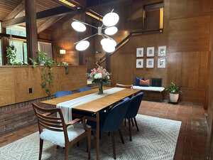 A dining room with a wooden table set for eight, blue and wooden chairs, a modern chandelier, a vase of flowers, wood-paneled walls, houseplants, framed art, and a bench with pillows and a fur throw on a patterned rug.