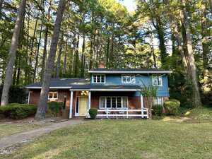A charming two-story house at 320 Granite Street in Henderson features a red brick lower level, blue siding above, tall pine trees and shrubs, a front lawn, and a welcoming white railing near the entrance.
