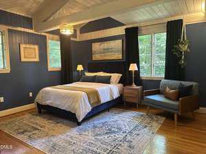 A bedroom with blue walls, a bed with white linens and beige throw, two bedside tables with lamps, a blue loveseat, large windows with dark curtains, a hanging plant, and a patterned area rug on a wooden floor.