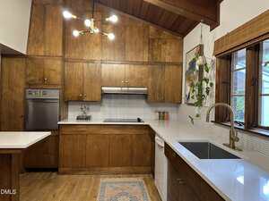 A kitchen with wood cabinets, a stainless steel oven, a stove with a range hood, white countertops, a large window with blinds, a sink with a gold faucet, a plant, and a modern light fixture.