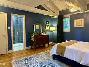 A bedroom with blue walls, a white ceiling with exposed beams, wooden floors, a bed with white and tan bedding, a blue patterned rug, a wooden dresser, a mirror, a plant, a lamp, and an open door to a bathroom.