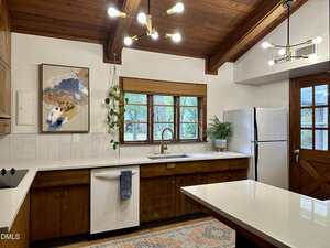 A kitchen with wood cabinets, white countertops, a white dishwasher and refrigerator, a large window with a bamboo shade, ceiling beams, two modern light fixtures, a hanging plant, a framed abstract painting, and a patterned rug.