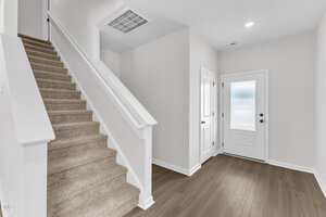 A modern entryway with light-colored walls, a white door with frosted glass, a small closet door, wood-style flooring, and a carpeted staircase with a white railing leading to the upper floor.
