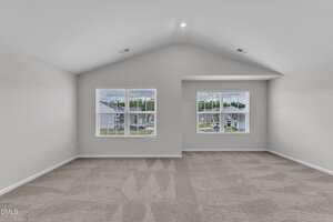 Spacious empty room with light gray walls, vaulted ceiling, and two large windows with white blinds letting in natural light. Beige carpet covers the floor, and suburban houses are visible outside.