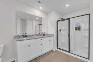A modern bathroom with a double-sink white vanity, granite countertop, large wall mirror, glass-enclosed shower with shelves, and wood-look flooring. The walls are white, and the lighting is recessed in the ceiling.