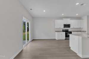 A modern kitchen with white cabinets, granite countertops, stainless steel appliances, and an open dining area. Sliding glass doors open to a grassy outdoor space. The room has light wood floors and white walls.