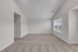 Empty room with light gray carpet, white walls, a vaulted ceiling, and two windows with blinds. Sunlight comes in through the windows, creating light patterns on the floor. The space appears clean and newly constructed.