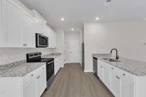 A modern kitchen with white cabinets, granite countertops, black appliances, a black faucet, and wood-style flooring. The room is brightly lit with recessed ceiling lights and has neutral-colored walls.
