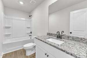 Modern bathroom with a white bathtub and shower, a toilet, and a vanity with a speckled granite countertop, rectangular sink, black faucet, and a large mirror. The floor is light wood, and the walls are white.