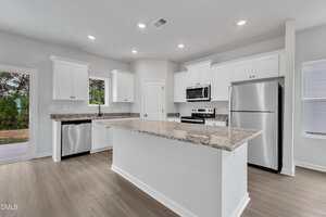 Modern kitchen with white cabinets, stainless steel appliances, granite countertops, a large central island, wood-style flooring, and recessed ceiling lights. A door and windows provide natural light and outdoor views.
