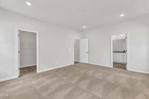 A spacious, empty bedroom with beige carpet, white walls, and three doorways leading to a walk-in closet, a bathroom with a double vanity, and a hallway. Recessed ceiling lights provide illumination.