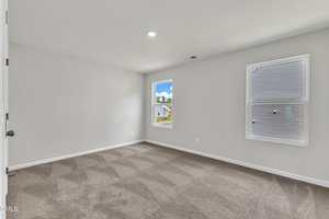 A clean, empty room with light gray walls, beige carpet, two windows with blinds, and a white ceiling with a single recessed light. Natural light comes in through the windows, and part of a house is visible outside.