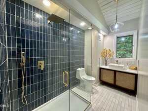 Modern bathroom with a glass-enclosed shower featuring blue tile, a white toilet, a wooden vanity with a vessel sink, a round mirror, a window, and a vase with yellow flowers on the counter.